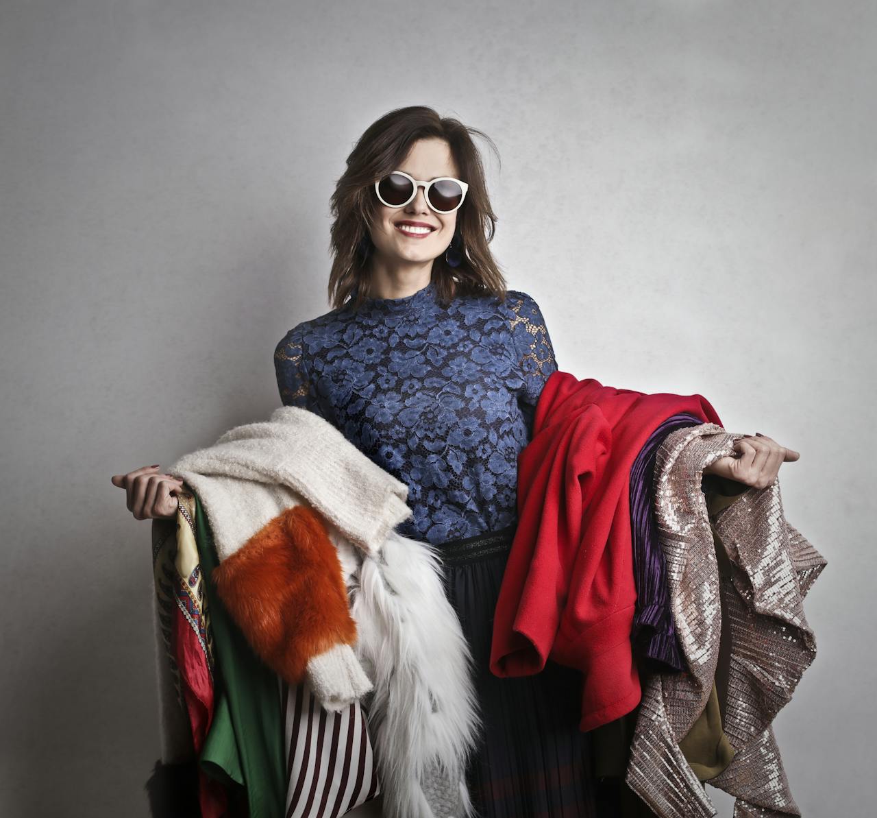 Elegant woman in sunglasses holding a variety of colorful outerwear, smiling at the camera.